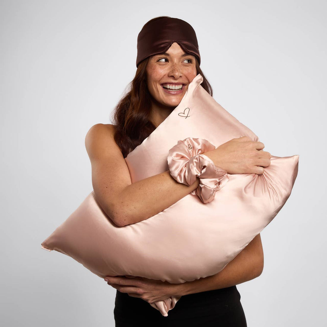 A Kitsch Nighttime Renewal 3pc Sleep Set with model with brown hair, holding in her arms a satin pillowcase, scrunchie on her wrist, and eye mask with heart details on her head.