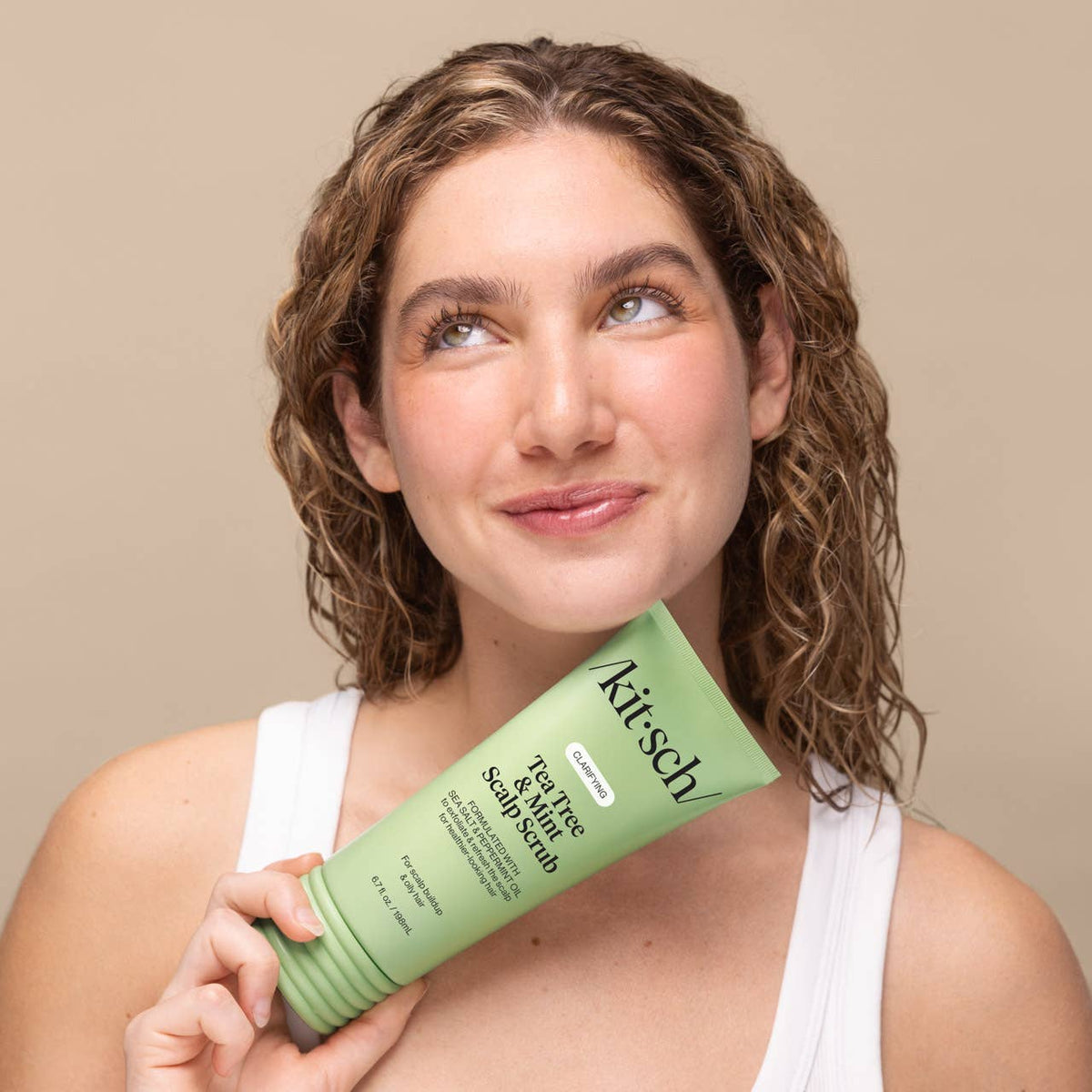 A woman with curly brown hair holds a tube of Kitsch Clarifying Tea Tree & Mint Scalp Scrub | Kiyo Beauty