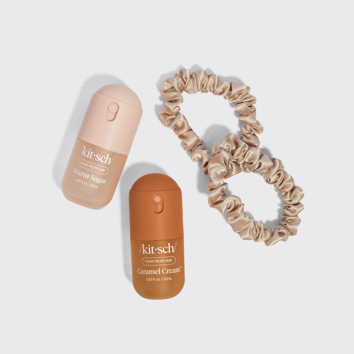 A flat lay of a beauty kit featuring two Kitsch liquid eyeshadow pots (Warm Sugar in a pale peach cap and Gimmel Cream in a brown cap) alongside two matching satin scrunchies in warm beige and tan shades, all arranged on a light gray background.