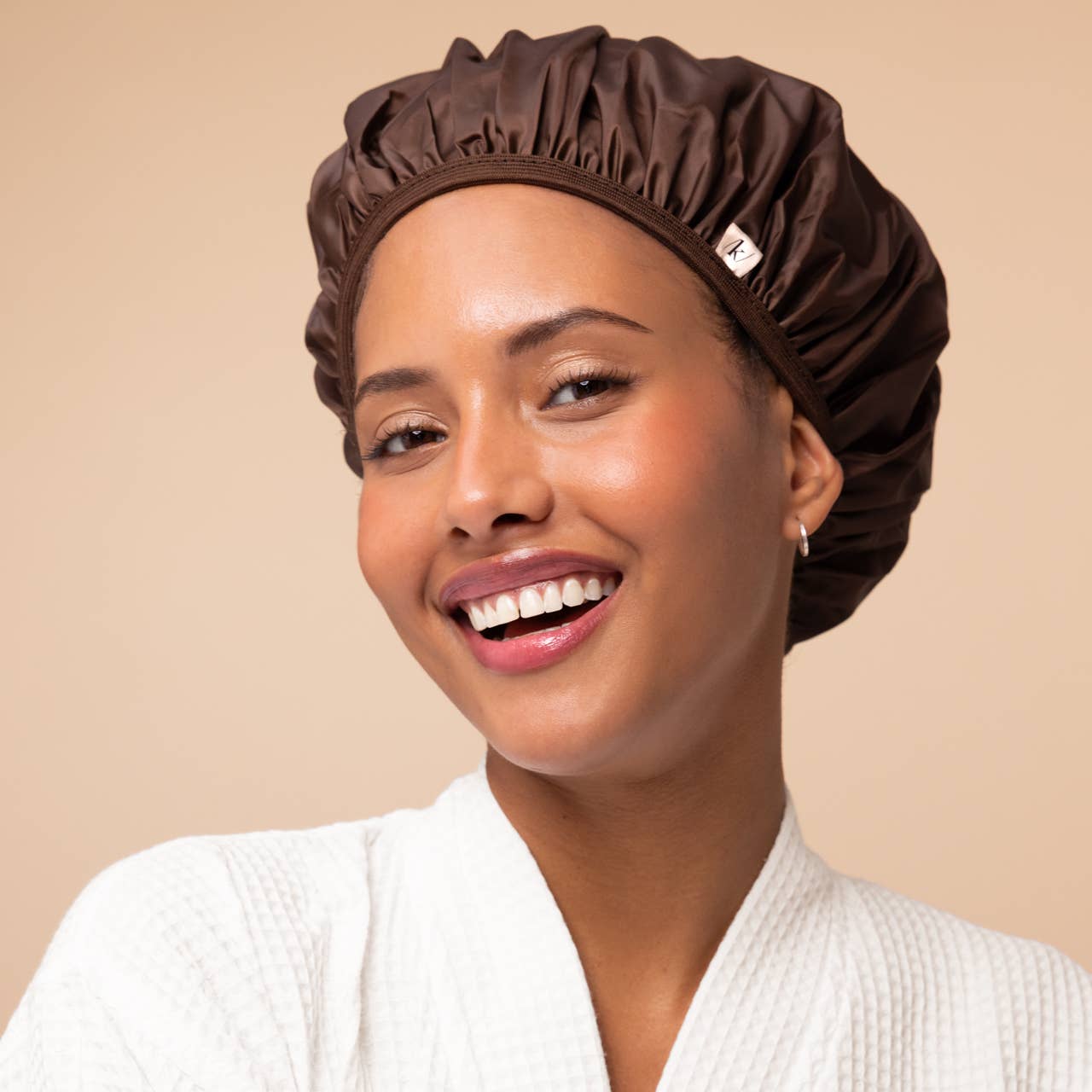 A smiling woman wearing a dark brown satin shower cap and a white textured robe against a solid beige background.