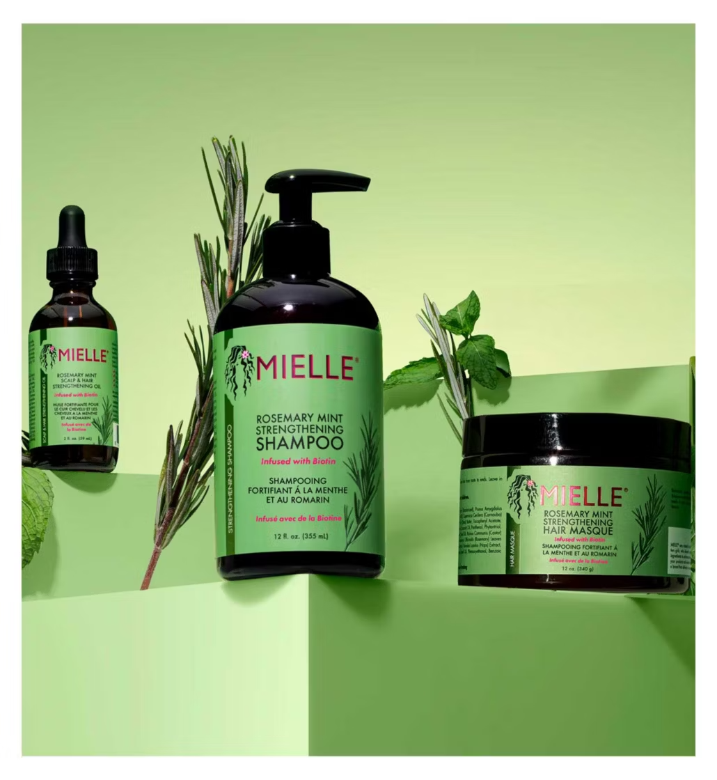 Three haircare products from the brand MIELLE are arranged on a light green background with botanical props: a small dropper bottle, a larger pump bottle of rosemary mint strengthening shampoo, and a jar of hair mask. The products feature dark amber/black packaging with bright green labels and pink brand lettering.