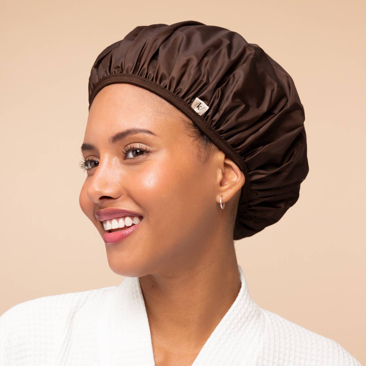 A smiling woman wearing a shiny dark brown satin shower cap and a white textured robe, shown in profile against a solid beige background.