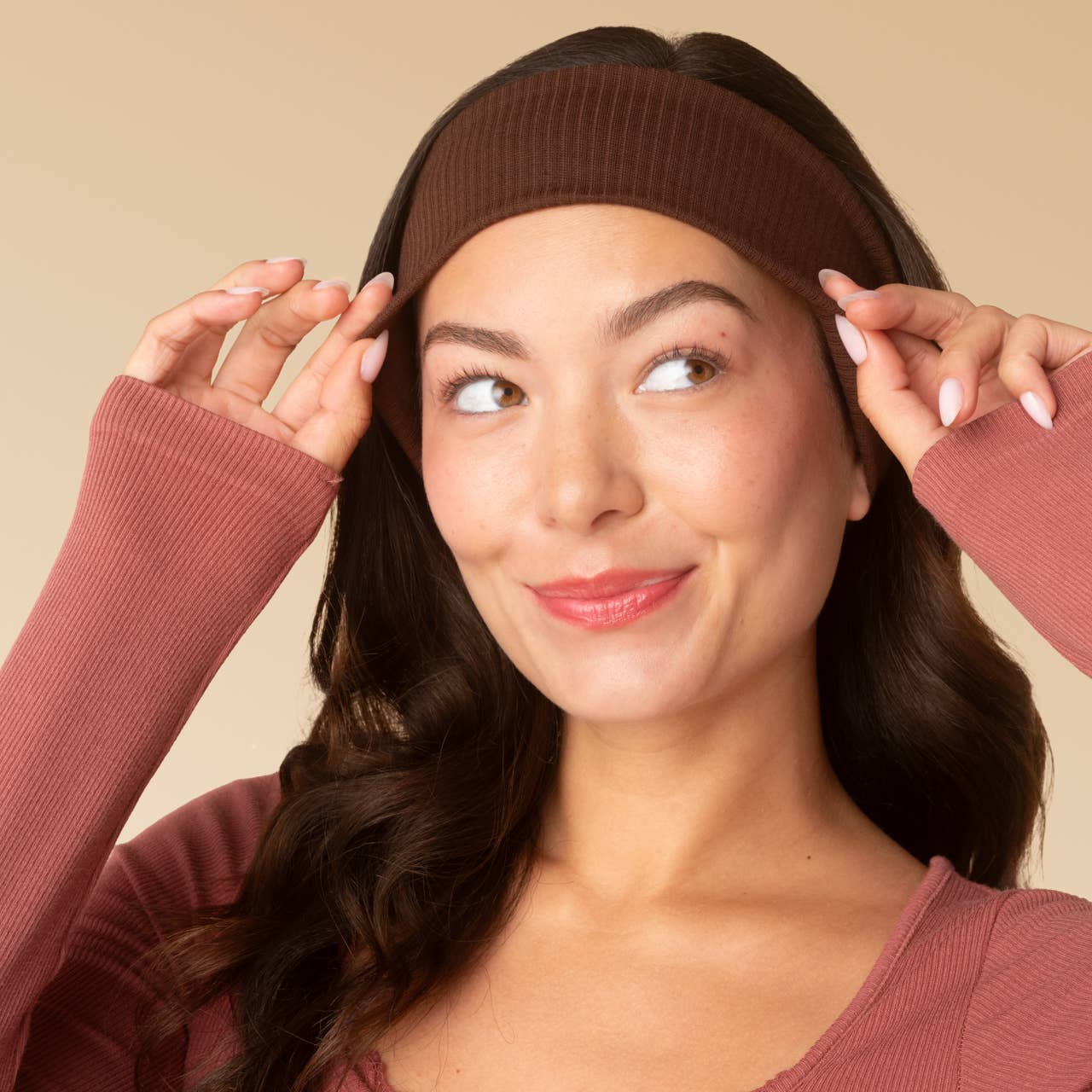 A woman wearing a ribbed brown headband and a matching ribbed long-sleeve top, posing with her hands on the sides of the headband against a solid beige background.