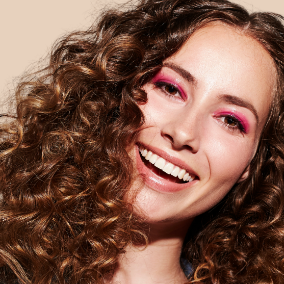 Woman with curly hair and pink makeup smiling against a beige background
