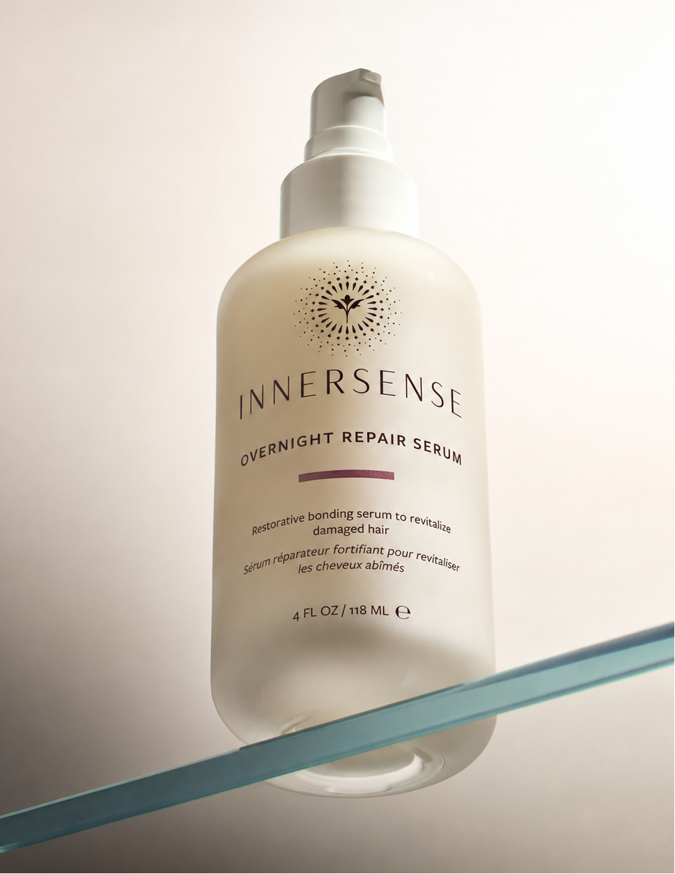 A frosted white bottle of Innersense Overnight Repair Serum with a white pump dispenser, resting on a clear glass shelf against a warm beige background.