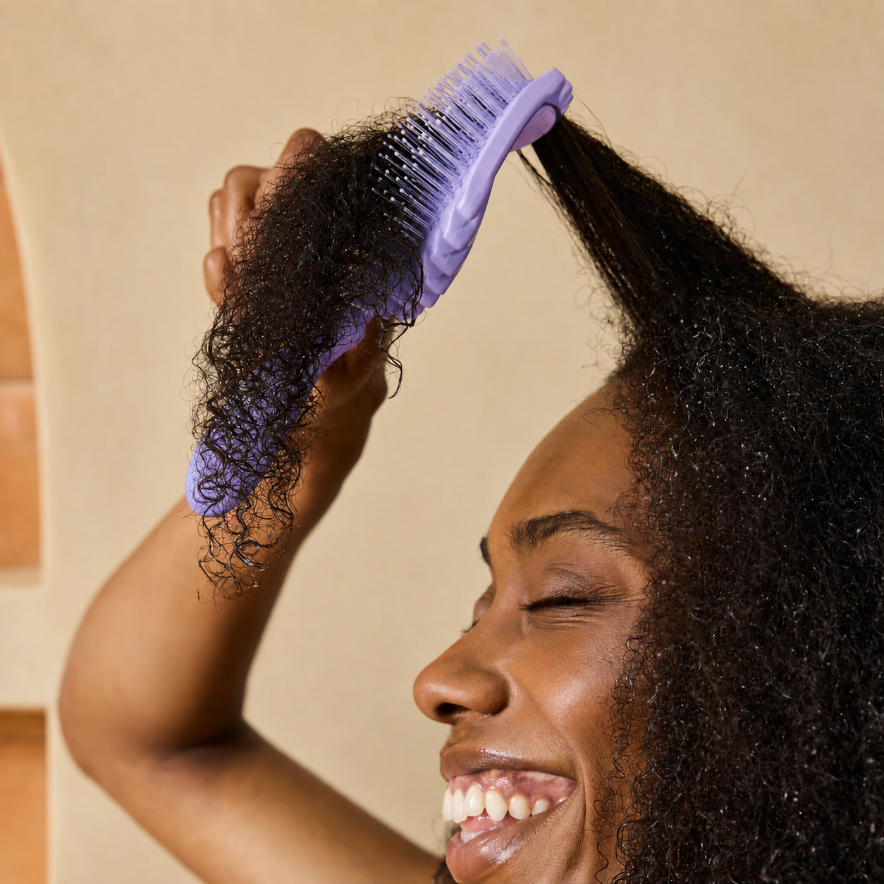 A close-up lifestyle shot of a woman with curly black hair smiling while using a lavender purple detangling brush on her hair, set against a warm beige background.