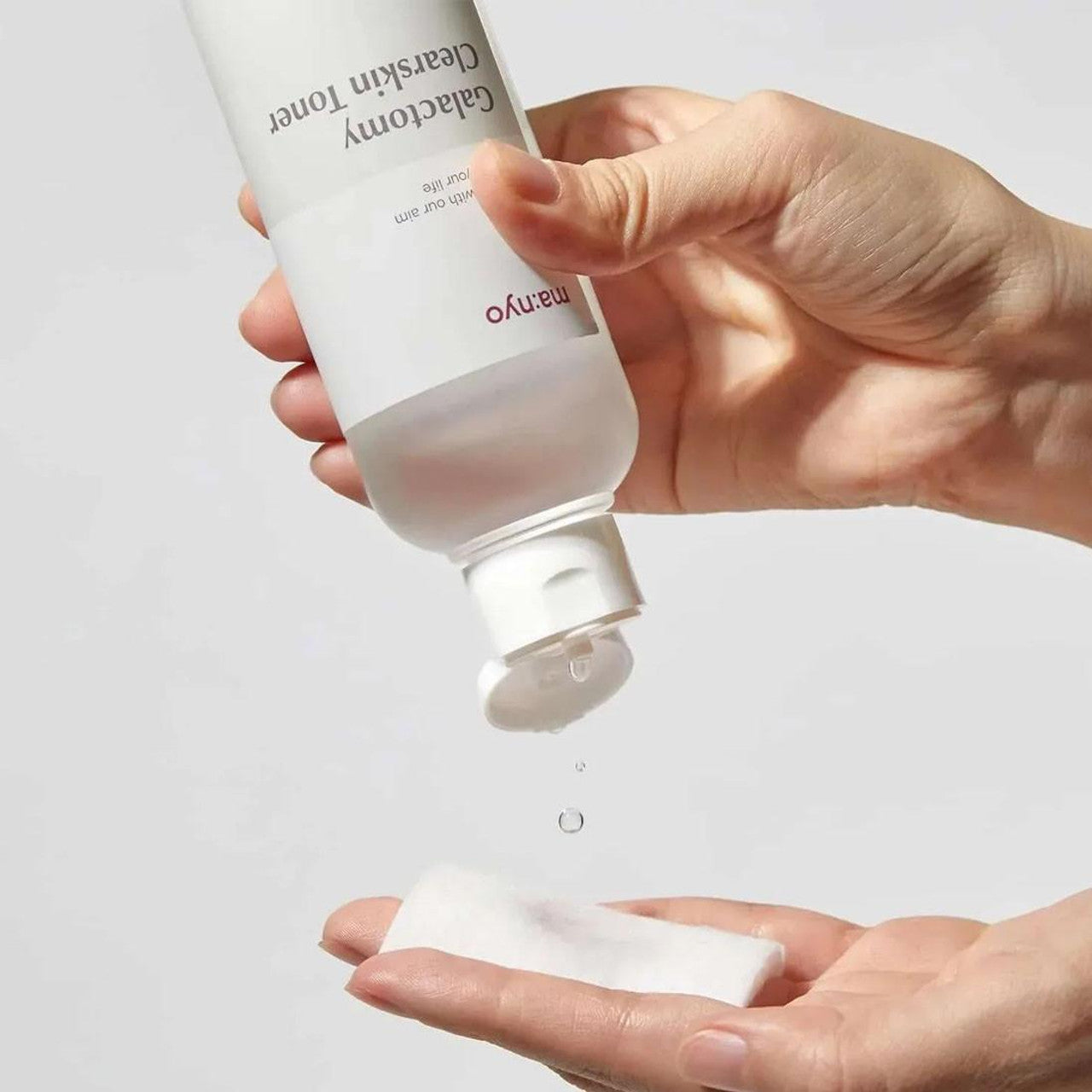 Hand holding a skincare bottle with a clear liquid being poured onto another hand against a light background