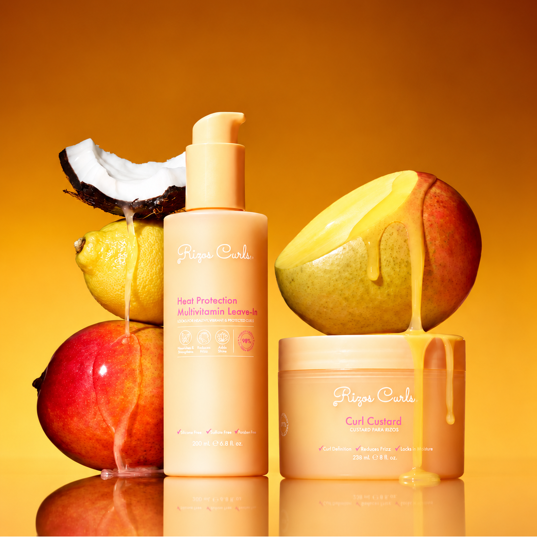 A set of Rizos Curls haircare products in a warm peach-orange color, styled with fresh fruit props including a whole mango, a sliced mango showing golden syrup, a lemon, and a coconut half, on a reflective surface against a warm orange gradient background.