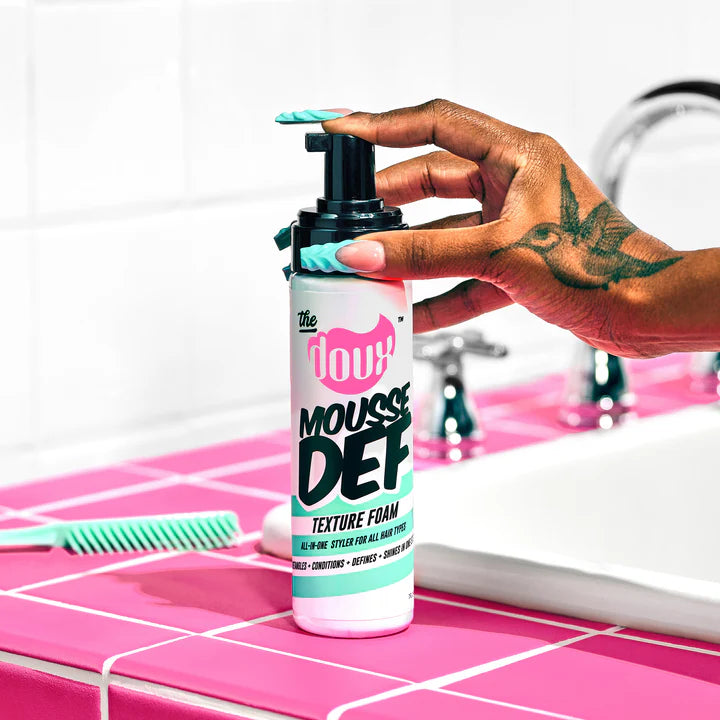 Hand holding a bottle of 'Mousse Def' texture foam on a pink tiled surface.